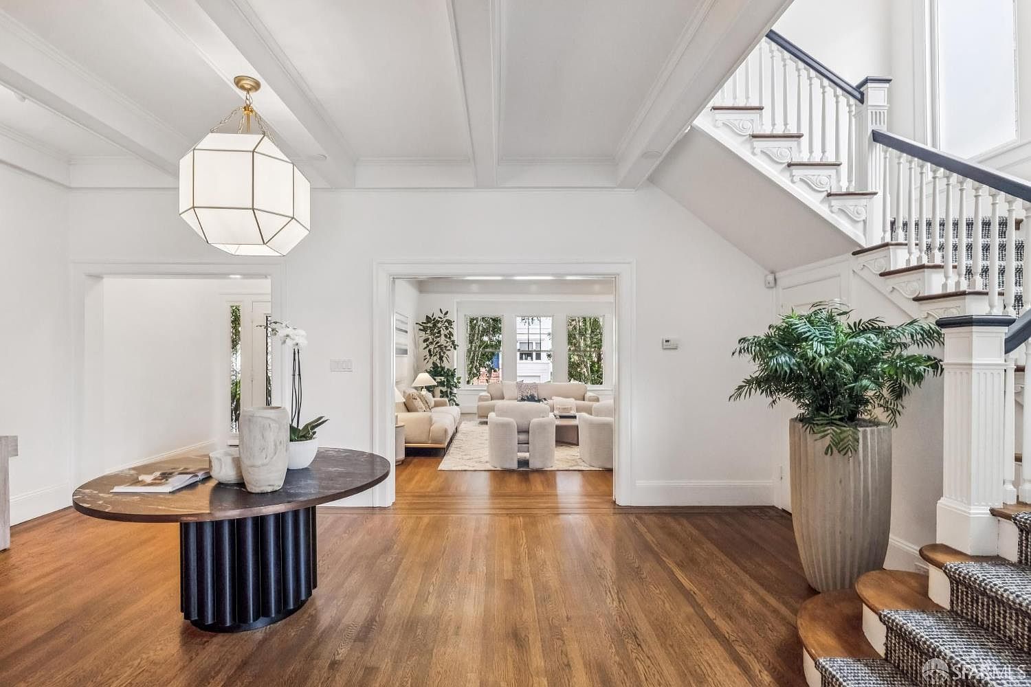 This is an interior shot of a grand foyer featuring hardwood floors, white walls, and a staircase with a dark railing. A round table with decorative vases sits to the left, while a large potted plant accents the base of the stairs. The space is bright and airy, creating a welcoming and elegant entrance.