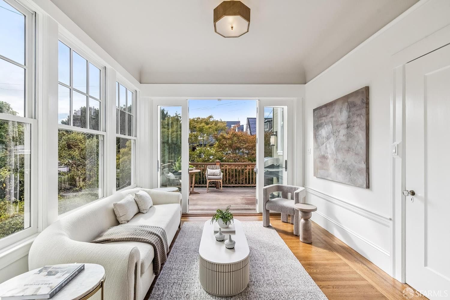 This is a bright and airy living room featuring a curved white sofa, a modern coffee table, and a neutral-toned rug. Large windows offer ample natural light and a view of the outdoors, while a sliding glass door leads to a balcony. The room is decorated with minimalist art and stylish furniture, creating a sophisticated and inviting atmosphere.