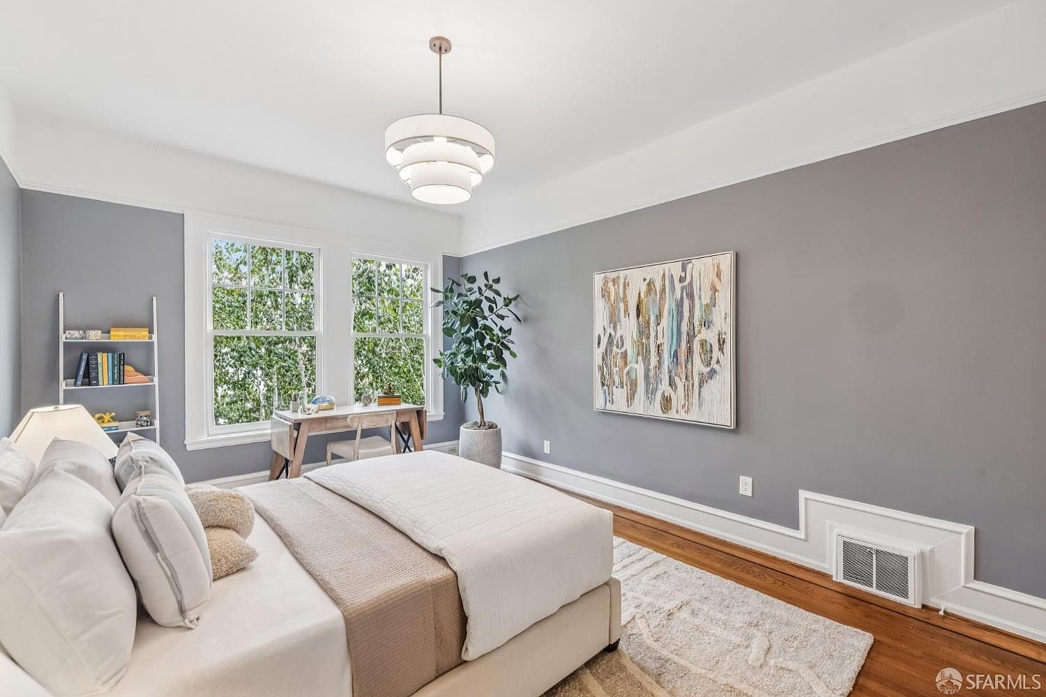 This is a bright and airy primary bedroom featuring a large bed with white linens and a beige throw, complemented by a neutral-toned rug. Natural light floods the room through two large windows, illuminating the gray walls and hardwood floors. A modern chandelier hangs from the ceiling, and artwork adorns the wall, creating a serene and inviting atmosphere.