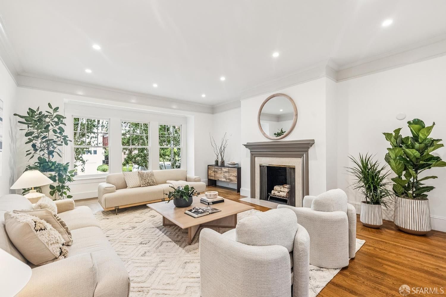 This is a bright and airy living room featuring hardwood floors, white walls, and recessed lighting. The room is furnished with two sofas, two armchairs, a coffee table, and a fireplace with a round mirror above it. Large windows provide natural light and views of the outdoors, creating a welcoming and comfortable atmosphere.
