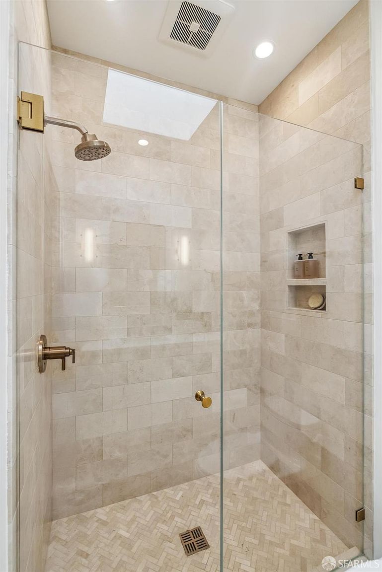 This is a well-lit primary bathroom shower featuring neutral-toned rectangular tiles and a glass enclosure. The shower includes bronze fixtures, a built-in niche for toiletries, and a herringbone-patterned tile floor. The perspective is from the entrance of the shower, showcasing the clean and modern design.