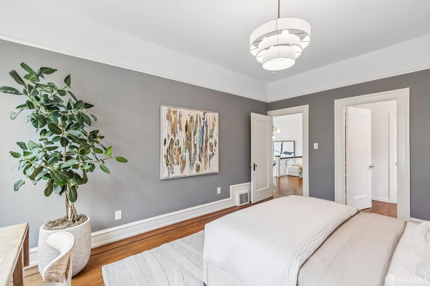 This is a primary bedroom featuring gray walls, hardwood floors, and white trim. A large abstract painting hangs on the wall, and a modern chandelier provides lighting. The room is furnished with a bed and a potted plant, creating a serene and inviting atmosphere.
