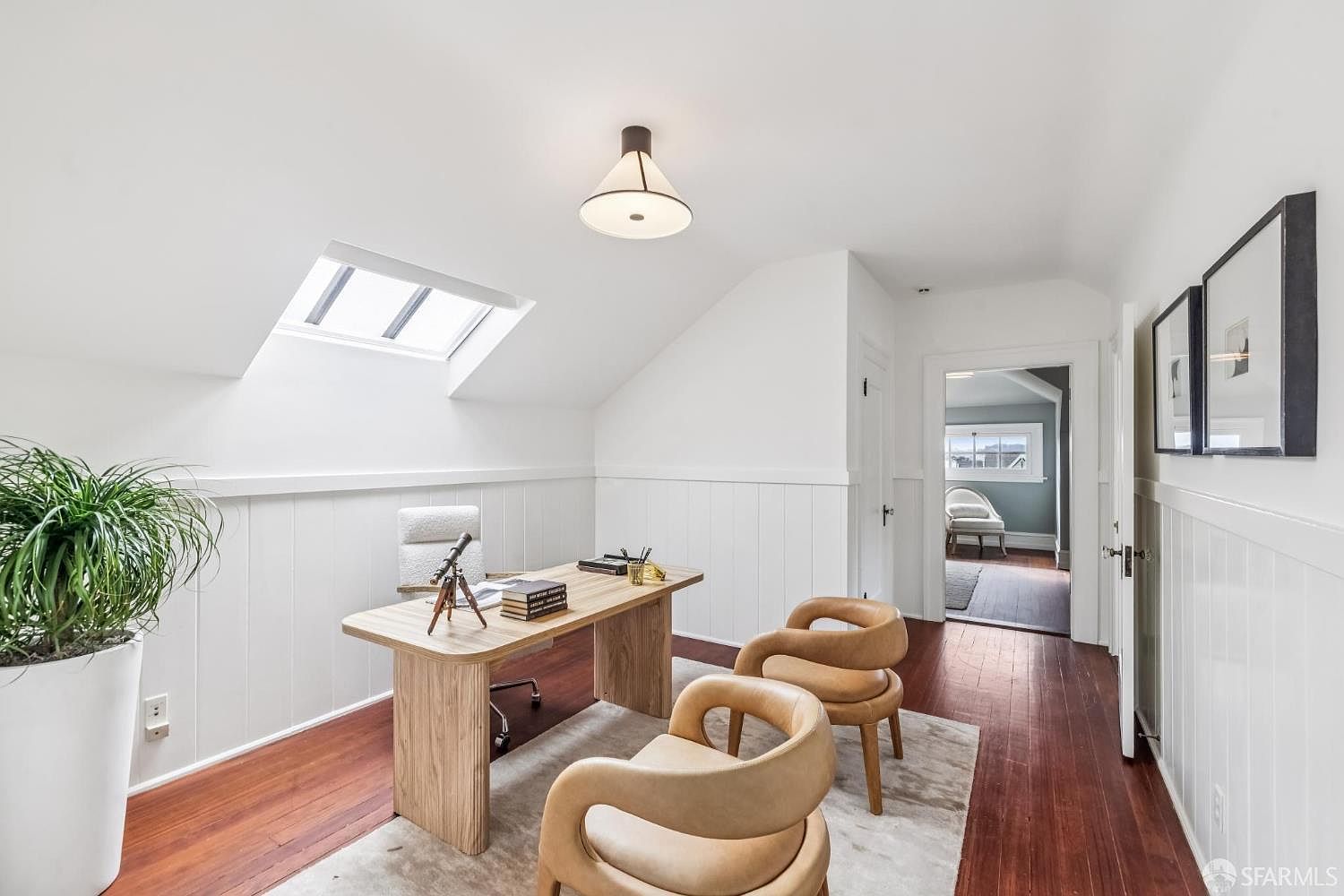 This is an interior shot of a bright and airy home office. The room features a wooden desk with a telescope and books, two stylish tan chairs, and a white rolling chair. Natural light floods the space through a skylight, and the walls are white with wainscoting, creating a clean and modern aesthetic.