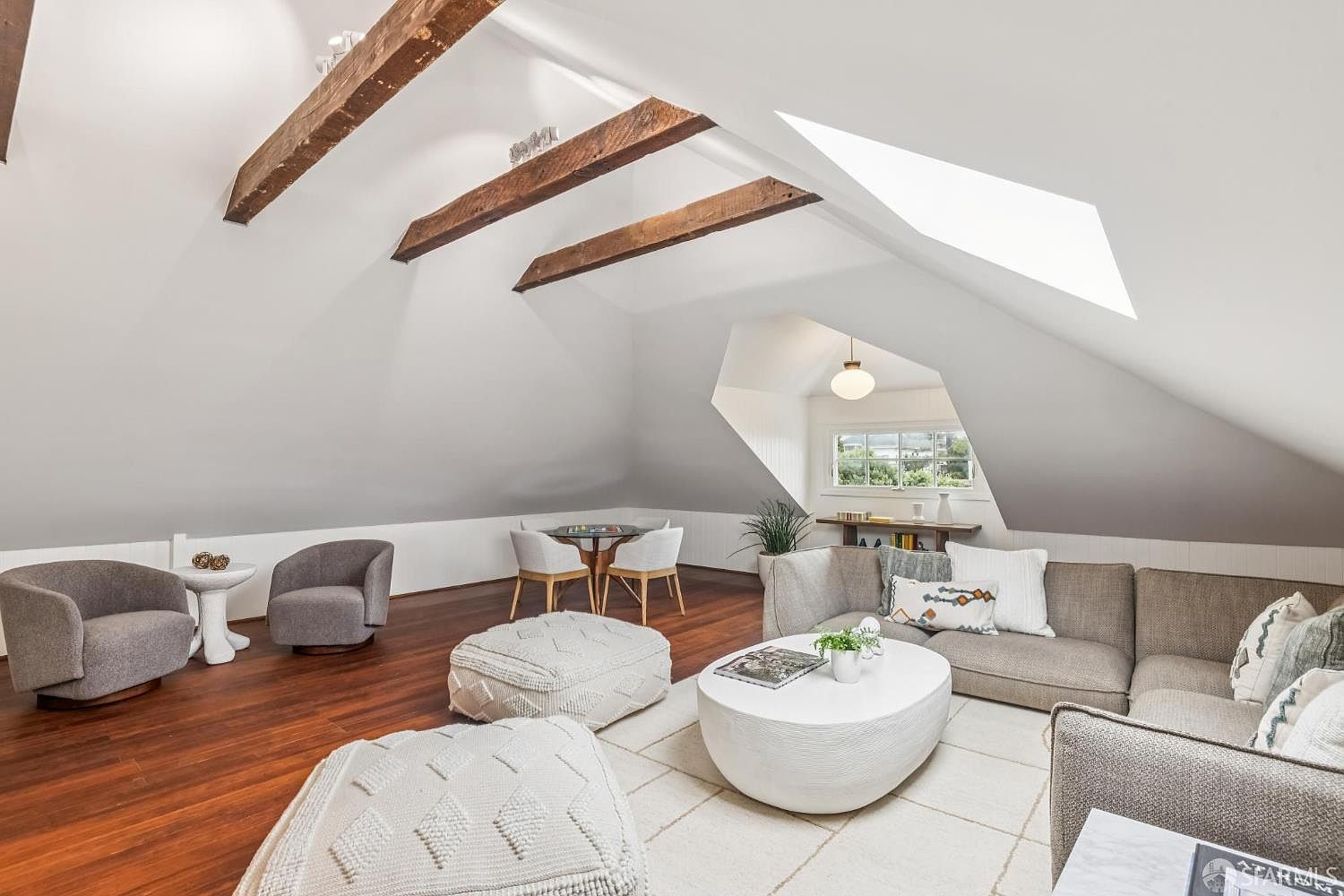 This is an interior shot of a living room in an attic space, featuring exposed wooden beams and a sloped ceiling with a skylight. The room is furnished with a sectional sofa, round coffee table, poufs, and armchairs, creating a cozy and inviting atmosphere. The hardwood floors and neutral color palette enhance the room's warmth and style.