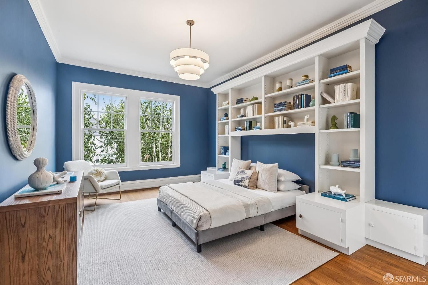 This is a beautifully designed primary bedroom featuring a built-in bookshelf headboard, painted in a striking blue hue. The room is well-lit with natural light from the windows, complemented by a modern chandelier. The hardwood floors and neutral-toned rug add warmth and comfort to the space, creating a serene and inviting atmosphere.