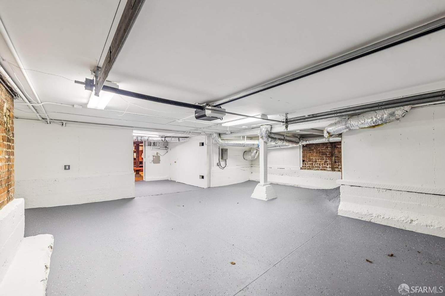 The image showcases a spacious basement with a gray epoxy floor and white-painted walls and ceiling. Exposed ductwork and pipes run along the ceiling, adding an industrial touch. A brick section is visible, providing a contrast to the otherwise modern, clean aesthetic. The room is well-lit, creating an open and airy feel.