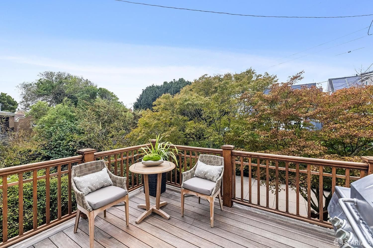 This image showcases a charming outdoor patio or deck area. Two wicker chairs with cushions flank a small round table adorned with a potted plant, creating an inviting space for relaxation. The wooden deck is surrounded by a railing, offering a view of lush greenery and trees, suggesting a private and serene setting.