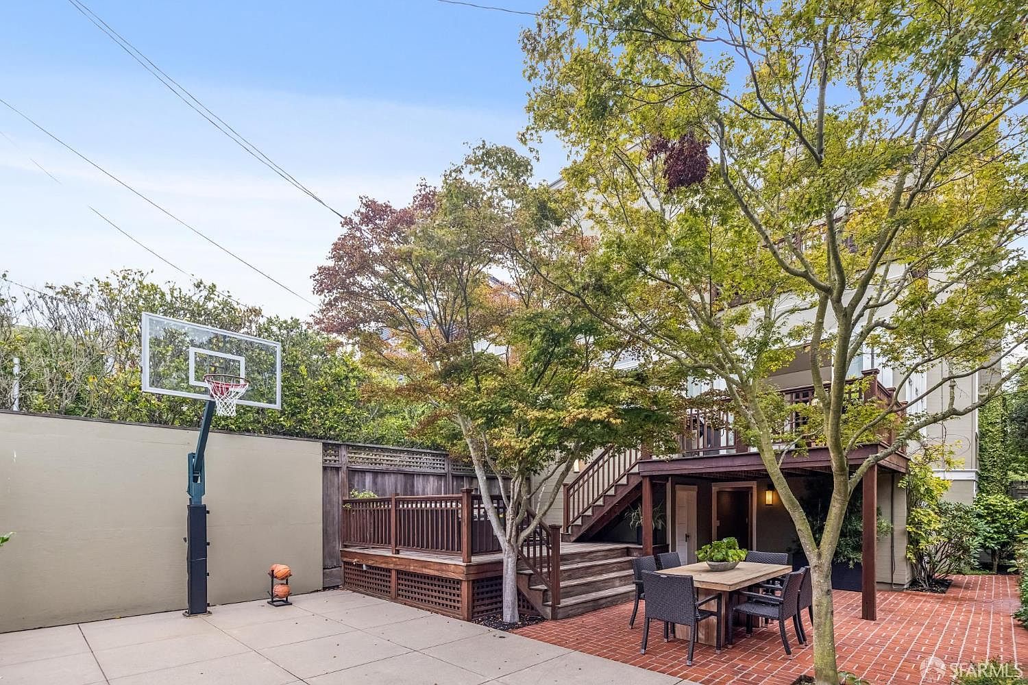 This exterior shot showcases a well-maintained backyard featuring a basketball hoop on a concrete slab, a wooden deck with stairs leading to an upper level, and a brick patio area with an outdoor dining set. Mature trees provide shade and privacy, enhancing the appeal of this outdoor living space. The overall impression is a functional and inviting backyard perfect for recreation and relaxation.
