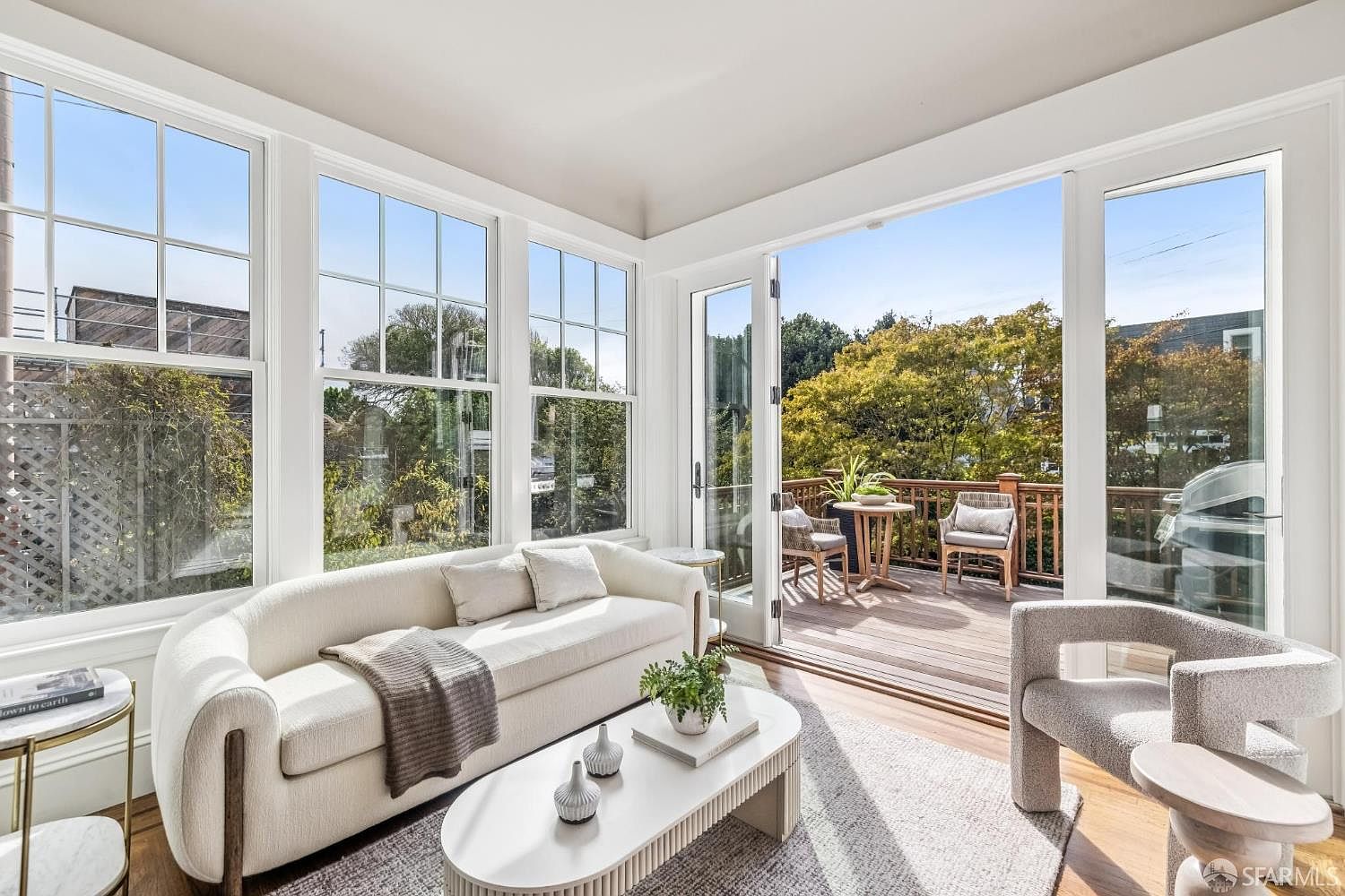 This is a bright and airy living room featuring a curved white sofa, a modern white coffee table, and a light-colored rug. Large windows and a glass door open to a deck, providing ample natural light and a seamless indoor-outdoor flow. The room is decorated in a minimalist style with neutral tones, creating a serene and inviting atmosphere.