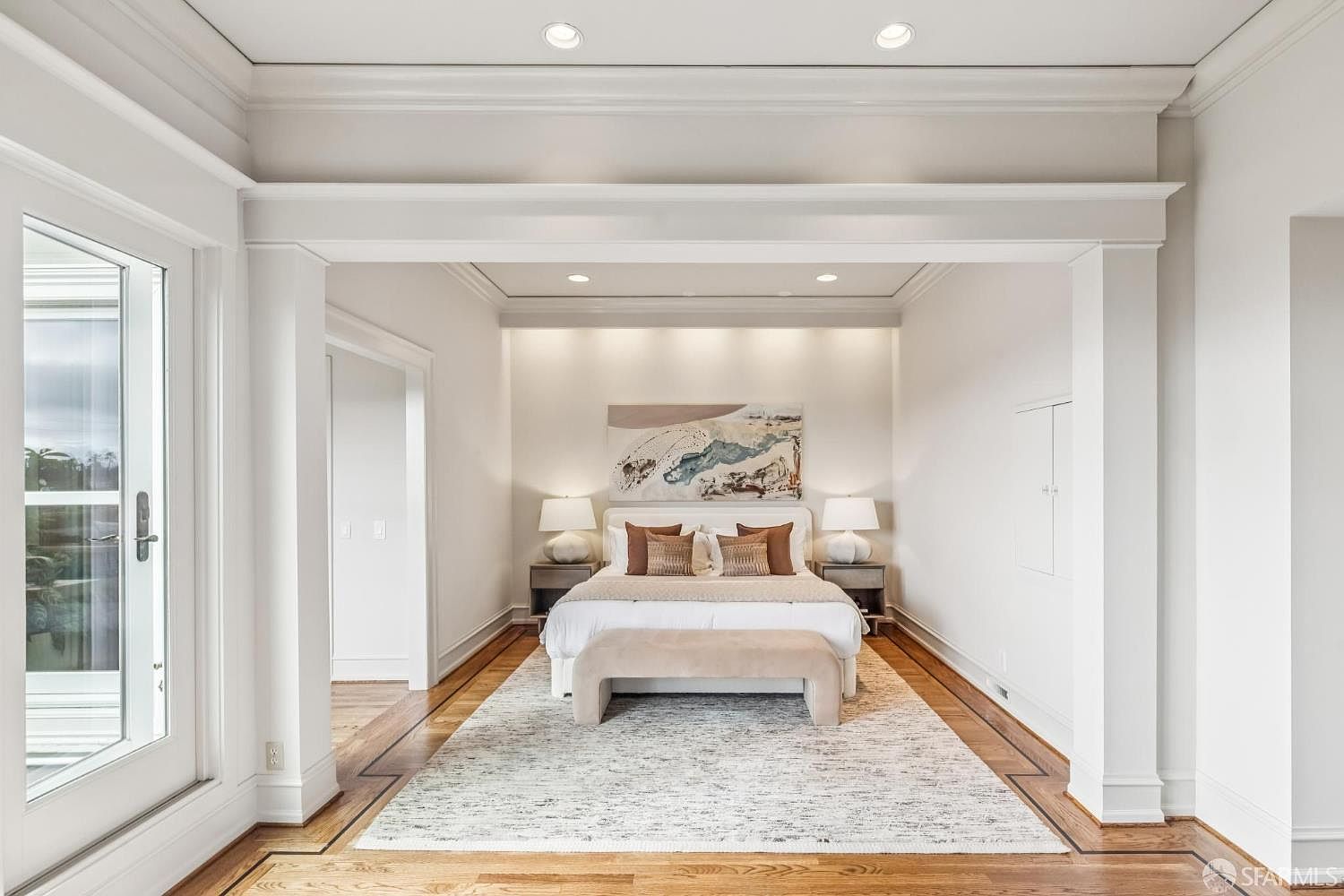 This is a bright and inviting primary bedroom featuring a neutral color palette. The room is well-lit with recessed lighting and natural light, highlighting the hardwood floors and a large area rug. A comfortable bed is centered in the room, flanked by nightstands and lamps, with an abstract painting above the headboard, creating a serene and luxurious atmosphere.