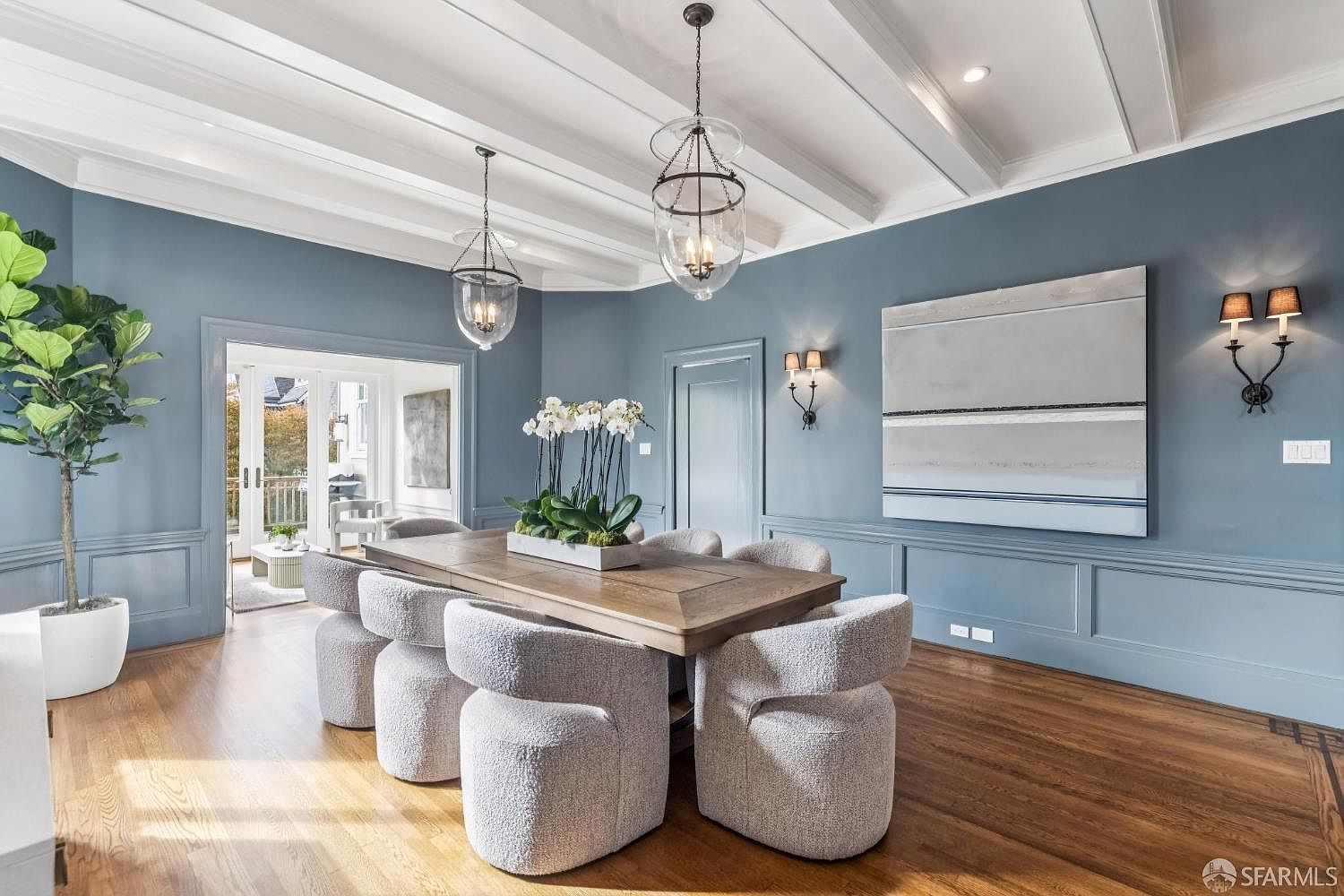 This is an interior shot of a dining room featuring a wooden table surrounded by six upholstered chairs. Two pendant lights hang above the table, and a large abstract painting adorns the wall. The room has a sophisticated and elegant feel, enhanced by the hardwood floors and wainscoting.