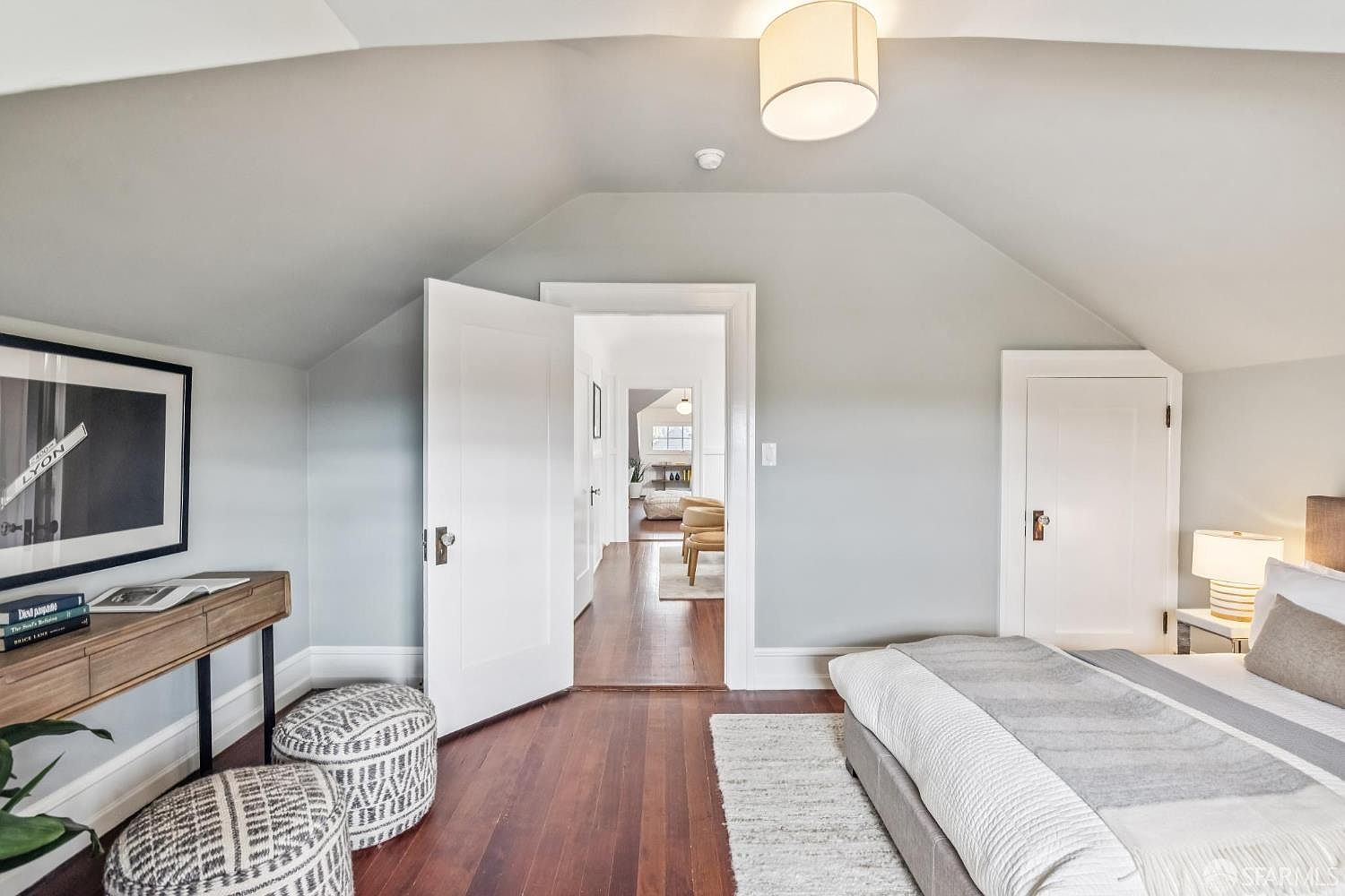 This is a bright and airy primary bedroom featuring hardwood floors, light gray walls, and white trim. The room includes a bed with a gray and white comforter, a wooden desk with a framed picture, and two patterned ottomans. An open doorway leads to another room, enhancing the sense of space and flow.