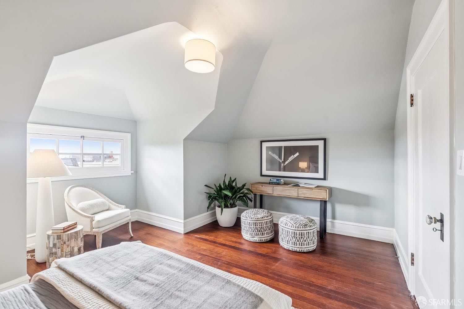 This is a cozy bedroom featuring hardwood floors, light gray walls, and an angled ceiling. The room is furnished with a white armchair, a small side table, a console table with a framed picture above it, and two decorative ottomans. Natural light streams in through a window, creating a bright and inviting atmosphere.