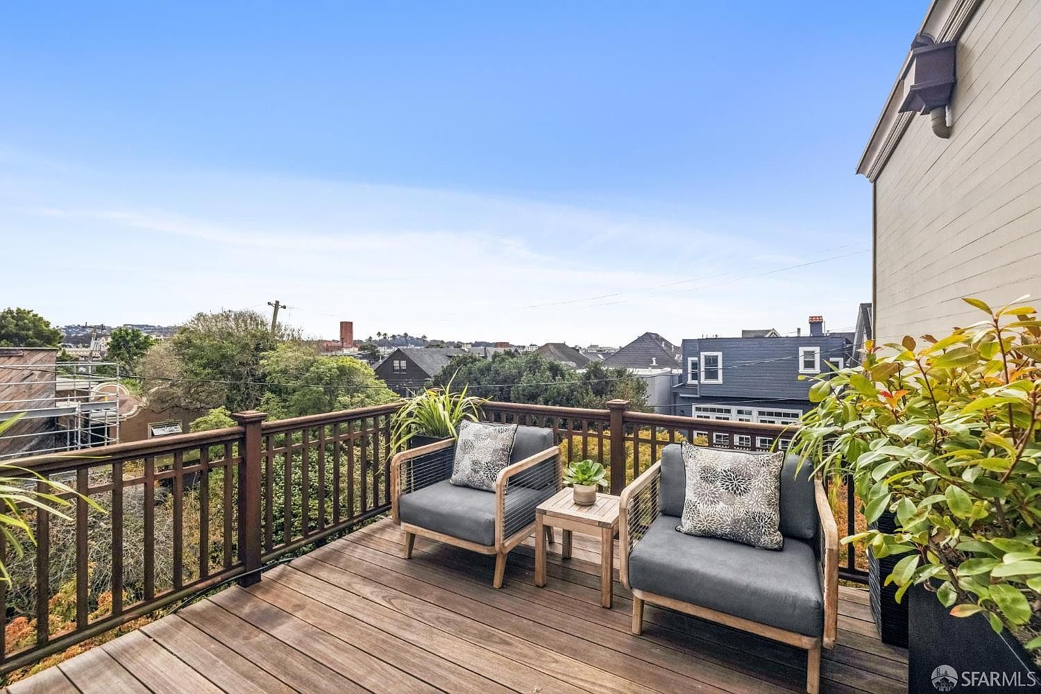 This image showcases a charming outdoor deck area, furnished with two modern armchairs and a small side table. The deck features wooden flooring and a railing, offering a pleasant view of the surrounding neighborhood and cityscape. Lush greenery in planters adds a touch of nature to the space, creating a relaxing and inviting atmosphere.