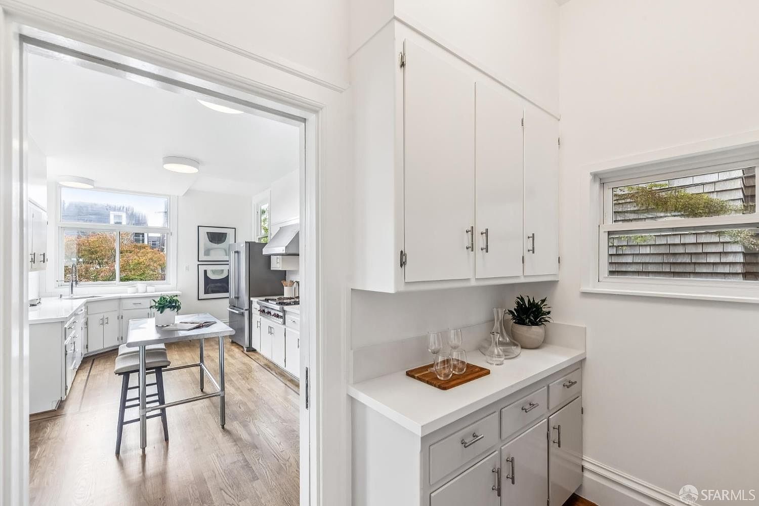 This interior shot showcases a bright, white kitchen with hardwood floors. The kitchen features white cabinetry with modern hardware, stainless steel appliances, and a small island with seating. A window provides natural light, and the overall impression is clean and inviting.