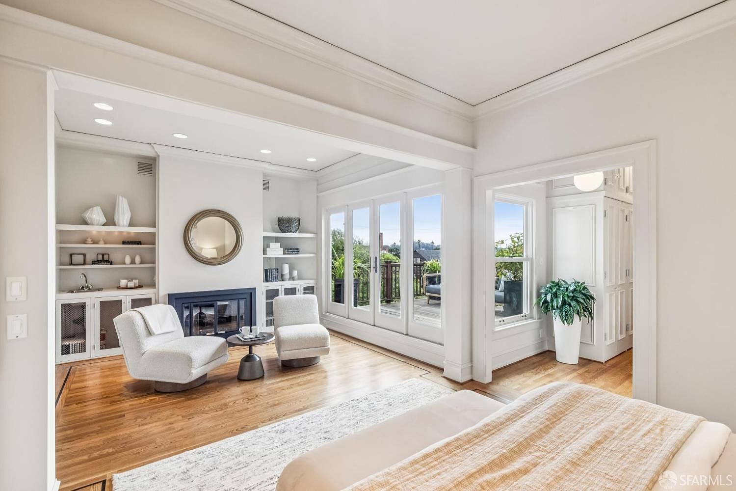 This is a bright and airy primary bedroom featuring a sitting area with two white armchairs flanking a fireplace, built-in shelving, and a round mirror. The room has hardwood floors, a light-colored area rug, and French doors leading to a balcony. The overall style is elegant and inviting, with a focus on natural light and comfortable living.