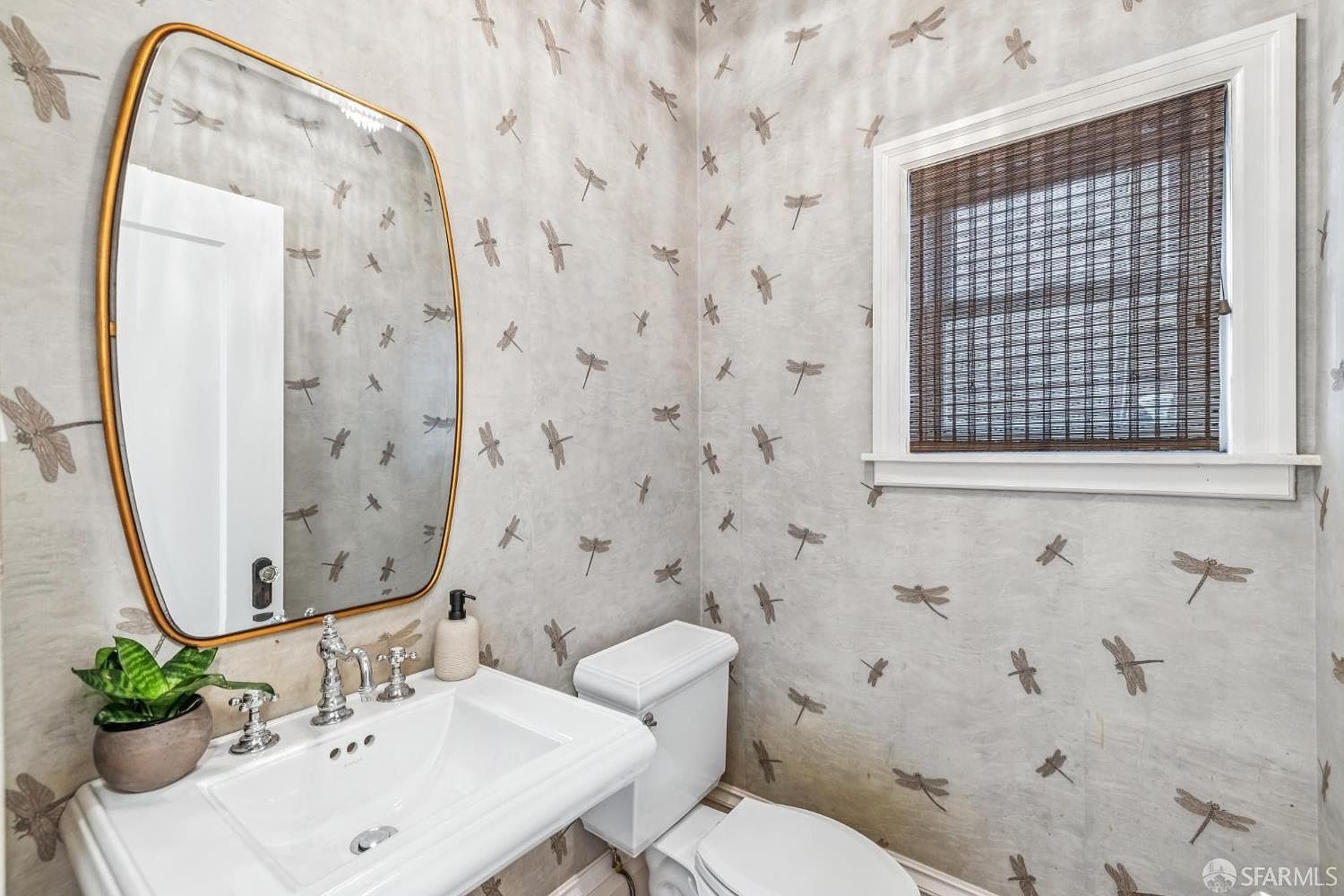 This is a well-decorated guest bathroom featuring dragonfly-patterned wallpaper, a white pedestal sink, and a matching toilet. A gold-framed mirror hangs above the sink, and a window with a woven shade provides natural light. The room has a charming and unique aesthetic.