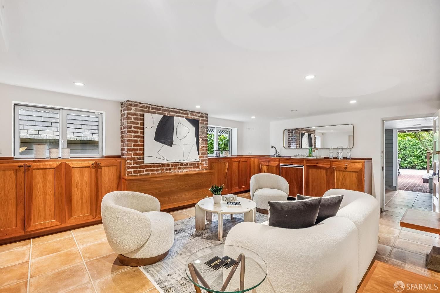 This interior shot showcases a stylish living room with a blend of textures and materials. The room features a brick accent wall with an abstract painting, warm wood cabinetry, and comfortable, rounded white seating arranged around a glass coffee table. The flooring is tiled, and the space is well-lit, creating an inviting and modern atmosphere.