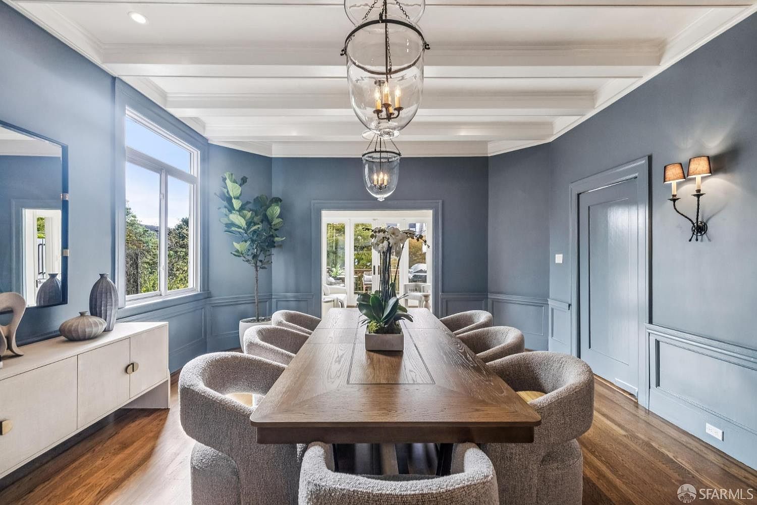 This is an interior shot of a dining room featuring a long wooden table surrounded by eight upholstered chairs. The room is painted in a muted blue, complemented by white trim and a coffered ceiling. A large window provides natural light, and a decorative light fixture hangs above the table, creating an elegant and inviting atmosphere.