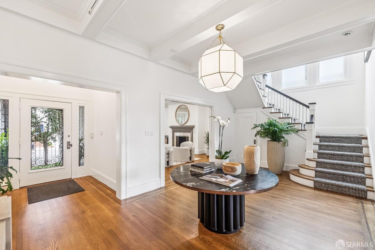 This is an interior shot of a grand entryway featuring hardwood floors, white walls, and a staircase with a patterned runner. A round table with decorative items and a large geometric pendant light add elegance to the space. The entryway leads to other rooms, creating a welcoming and sophisticated first impression.
