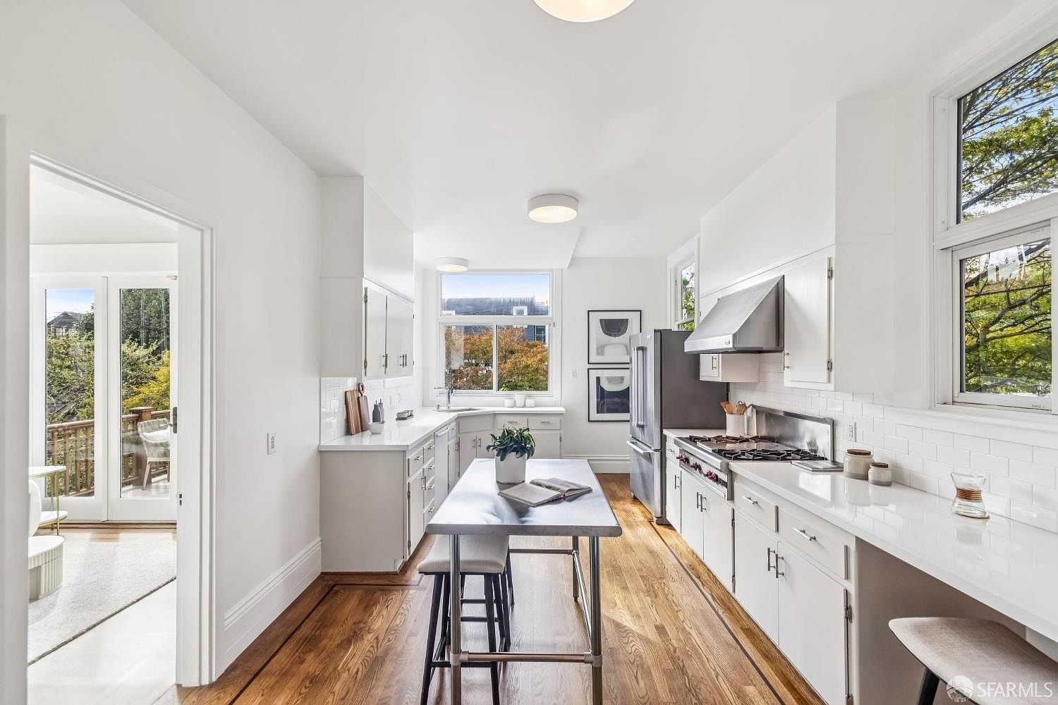 This is a bright and airy kitchen featuring white cabinetry, countertops, and walls, complemented by warm hardwood flooring. A central island with seating provides a focal point, while stainless steel appliances and a window overlooking greenery add modern touches. The kitchen's design emphasizes clean lines and natural light, creating an inviting and functional space.