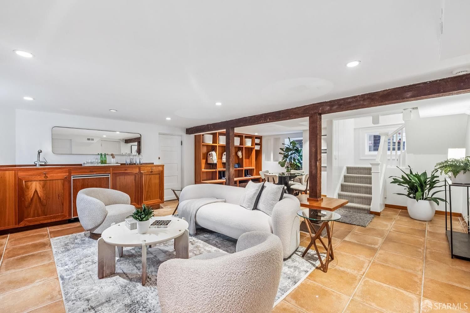 This is an interior shot of a living room featuring a curved white sofa, two armchairs, and a round coffee table on a patterned rug. The room has a built-in wooden bar area, exposed wooden beams, and a staircase leading to another level. The flooring is tile, and the overall style is modern with rustic touches.