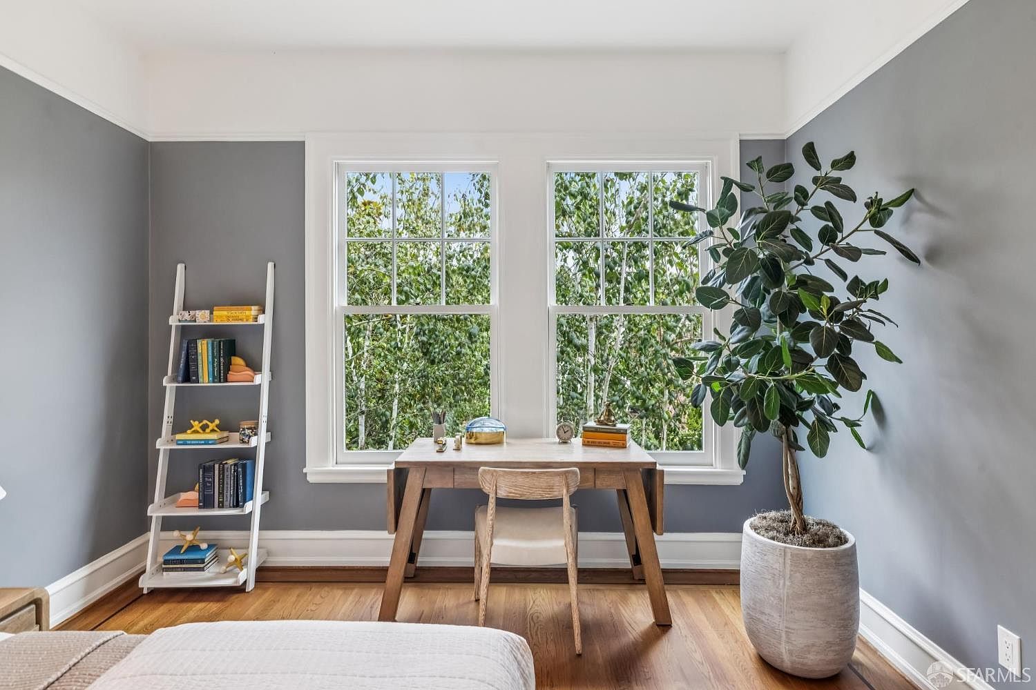 This is an interior shot of a home office or study area. The room features a wooden desk positioned in front of two windows that offer a view of lush greenery. A white ladder bookshelf stands against the wall, and a large potted plant adds a touch of nature to the space. The walls are painted a neutral gray, creating a calm and inviting atmosphere.