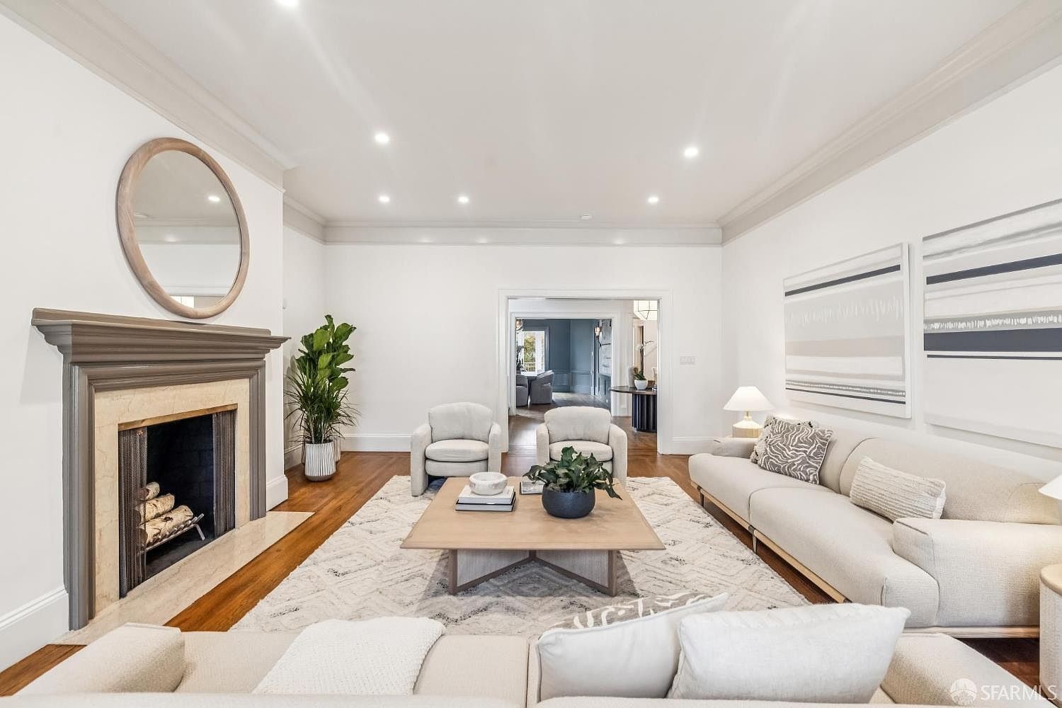 This is a bright and airy living room featuring a neutral color palette. The room is furnished with two armchairs, a sofa, and a coffee table, all arranged on a patterned area rug. A fireplace with a round mirror above it serves as a focal point, and artwork adorns the walls. The perspective is from the front of the room, looking towards the fireplace and seating area.