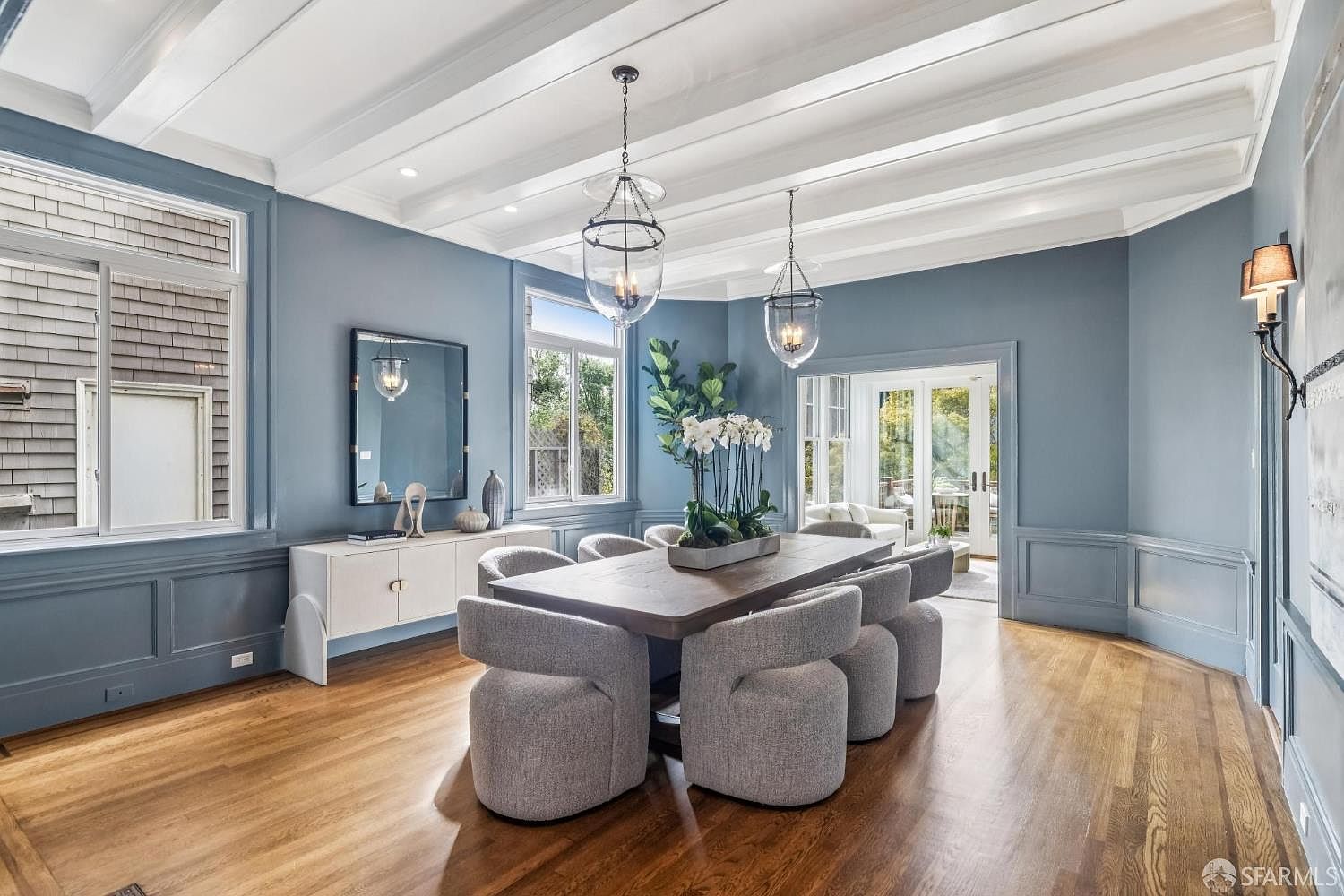 This is an interior shot of a dining room featuring a large wooden table surrounded by eight gray upholstered chairs. Two modern pendant lights hang above the table, and a sideboard with decorative items sits against one wall. The room is painted in a muted blue, with white trim and hardwood floors, creating an elegant and inviting atmosphere.