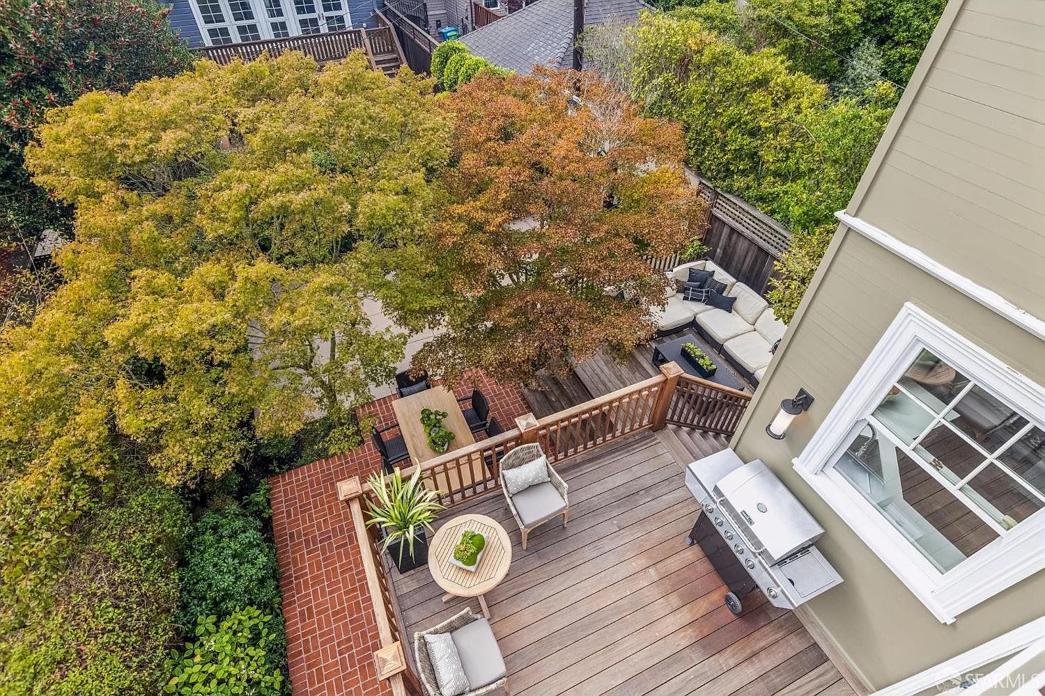 This is an aerial view of a well-appointed outdoor deck and patio area. The space features a dining table with chairs, a lounge area with a sectional sofa, and a grilling station with a stainless steel grill. The deck is surrounded by lush trees and greenery, creating a private and inviting outdoor living space.