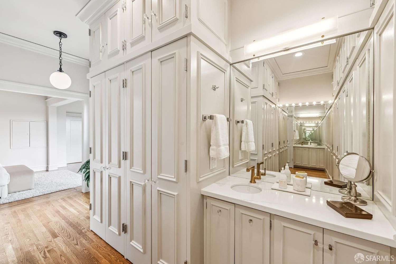 This is a well-organized primary bathroom featuring custom white cabinetry with detailed trim and crystal hardware. A large mirror reflects the length of the room, creating an illusion of depth and showcasing the extensive storage. The vanity area includes a white countertop, gold-toned fixtures, and decorative accessories, contributing to a luxurious and elegant atmosphere.