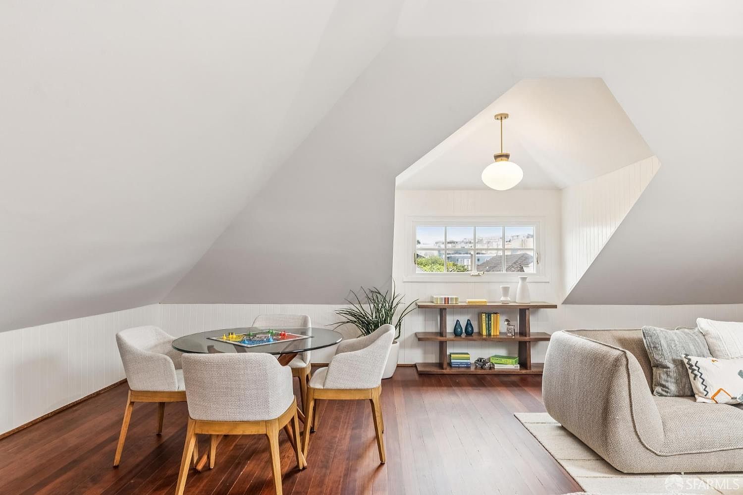 This interior shot showcases a charming attic space converted into a functional living area. The room features a pitched roof, hardwood floors, and a cozy seating arrangement with a round glass table and four chairs. A built-in bookshelf and a window offering a glimpse of the neighborhood add to the room's appeal, creating a warm and inviting atmosphere.