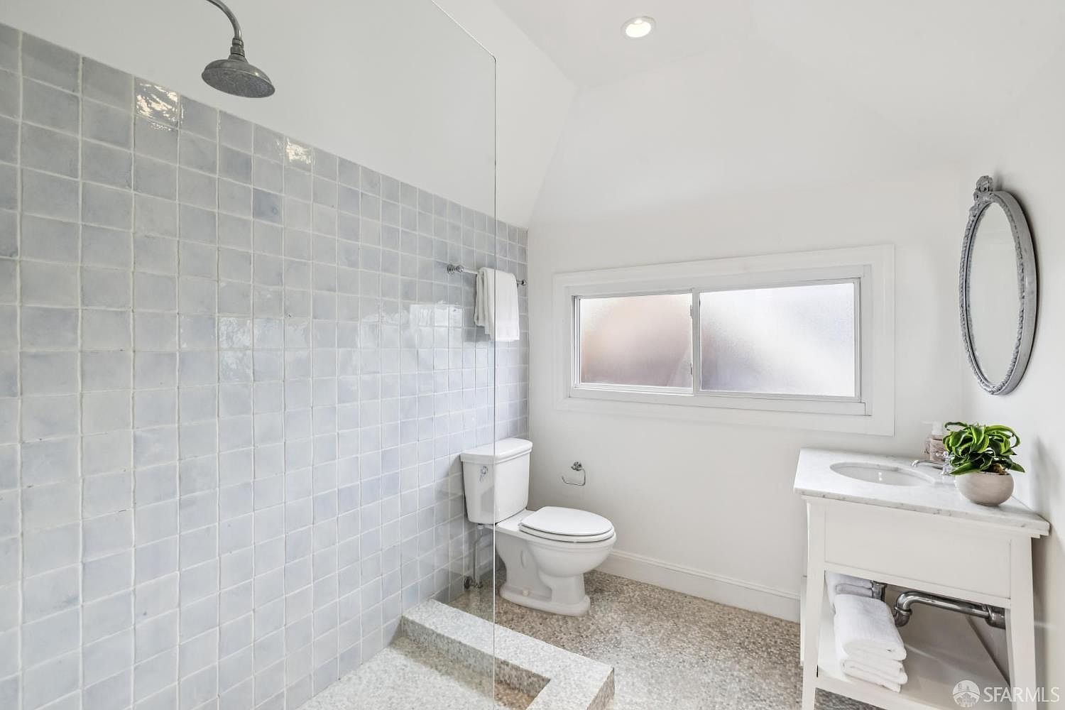This is a bright and clean bathroom featuring a walk-in shower with light blue square tiles and a glass partition. The room includes a white toilet, a vanity with a marble countertop, and an oval mirror. A small plant adds a touch of nature, and the overall aesthetic is simple and modern.