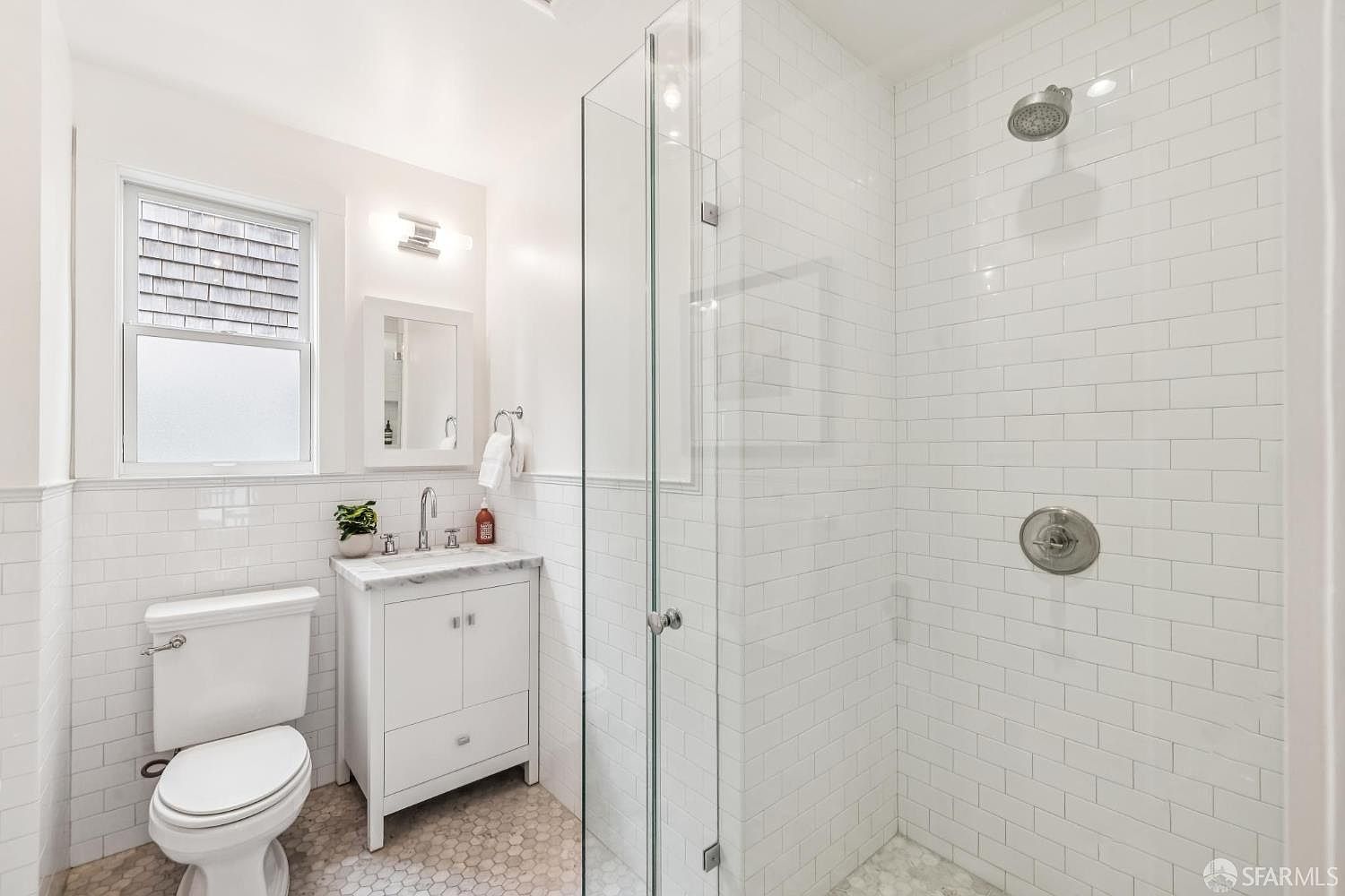 This is a well-lit bathroom featuring white subway tile on the walls and hexagonal tile flooring. A white vanity with a marble countertop is positioned next to a toilet, and a glass-enclosed shower is visible on the right. The overall impression is clean, bright, and modern.