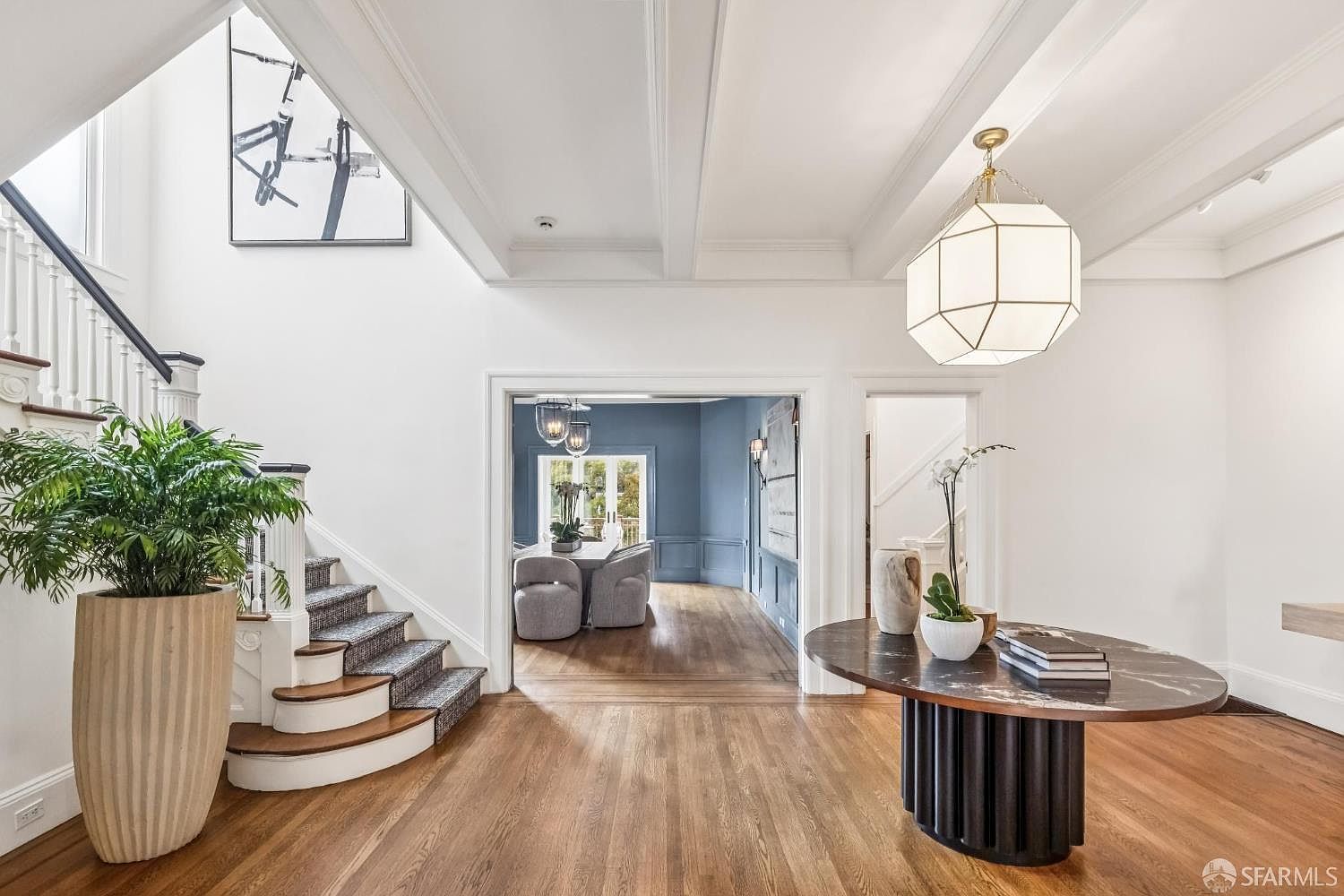 This is an interior shot of a grand hallway featuring a staircase, a round table with decorative items, and a large potted plant. The walls are painted white, and the hardwood floors add warmth to the space. The lighting is provided by a modern chandelier, and an open doorway leads to a dining area with blue walls.