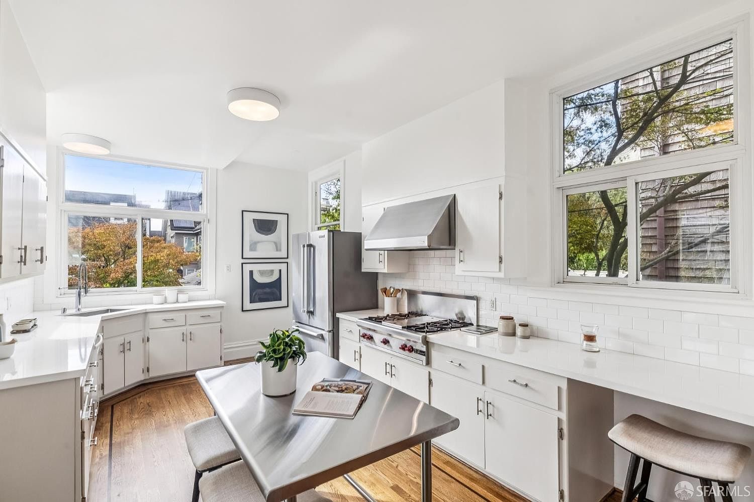 This is a bright and airy kitchen featuring white cabinetry, stainless steel appliances, and hardwood floors. Natural light floods the space through large windows, offering pleasant views of the outdoors. A stainless steel table with seating provides a modern touch, while the white subway tile backsplash adds a classic element.