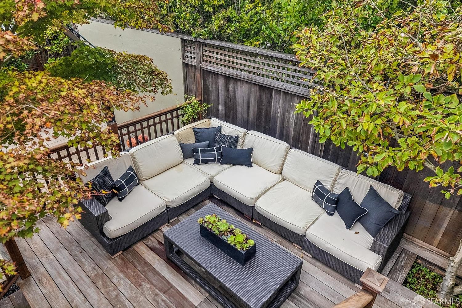 This is a high-angle shot of a cozy outdoor patio area. The space features a large, sectional sofa with cream cushions and dark throw pillows, arranged around a dark wicker coffee table with a succulent centerpiece. The patio is surrounded by a wooden fence and lush greenery, creating a private and inviting outdoor living space.