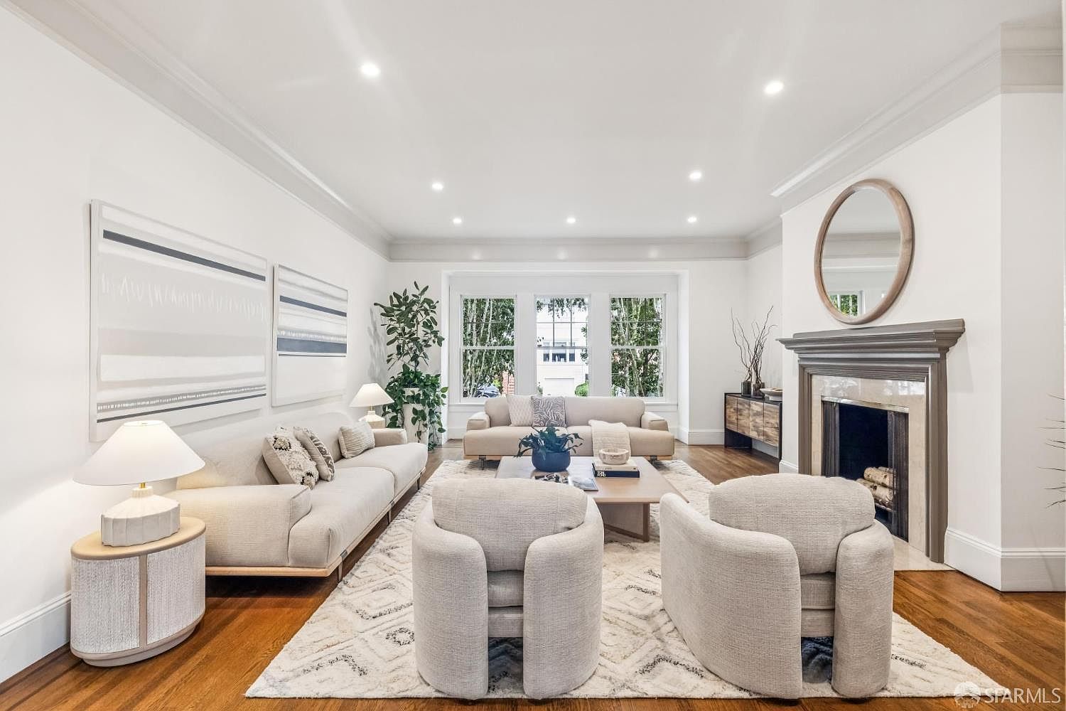 This is a bright and spacious living room featuring hardwood floors, white walls, and recessed lighting. The room is furnished with two sofas, two armchairs, a coffee table, and a fireplace with a round mirror above it. Large windows provide natural light, and artwork adorns the walls, creating a welcoming and elegant atmosphere.