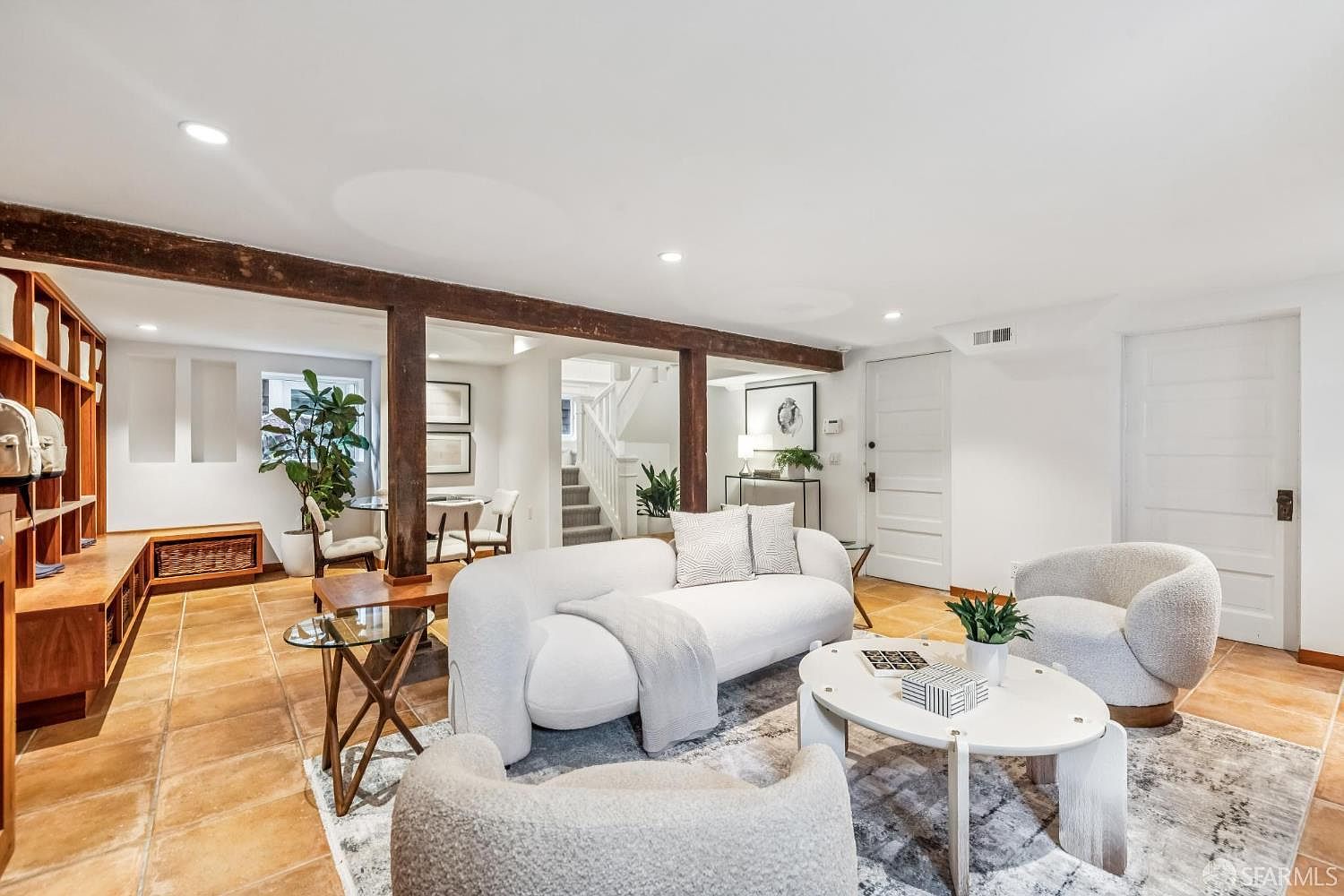 This is an interior shot of a living room featuring a white sofa, two armchairs, and a round coffee table on a patterned rug. The room has exposed wooden beams and a built-in shelving unit, creating a cozy and inviting atmosphere. The flooring is tile, and the walls are painted white, providing a bright and clean aesthetic.