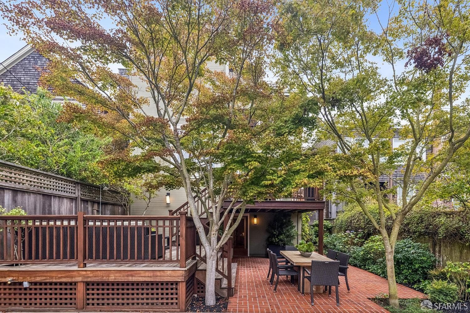 This image showcases a charming backyard patio and deck area. The patio is paved with red bricks and features an outdoor dining set, while a wooden deck with railings offers additional space for relaxation. Mature trees provide shade and privacy, enhancing the appeal of this outdoor living space.