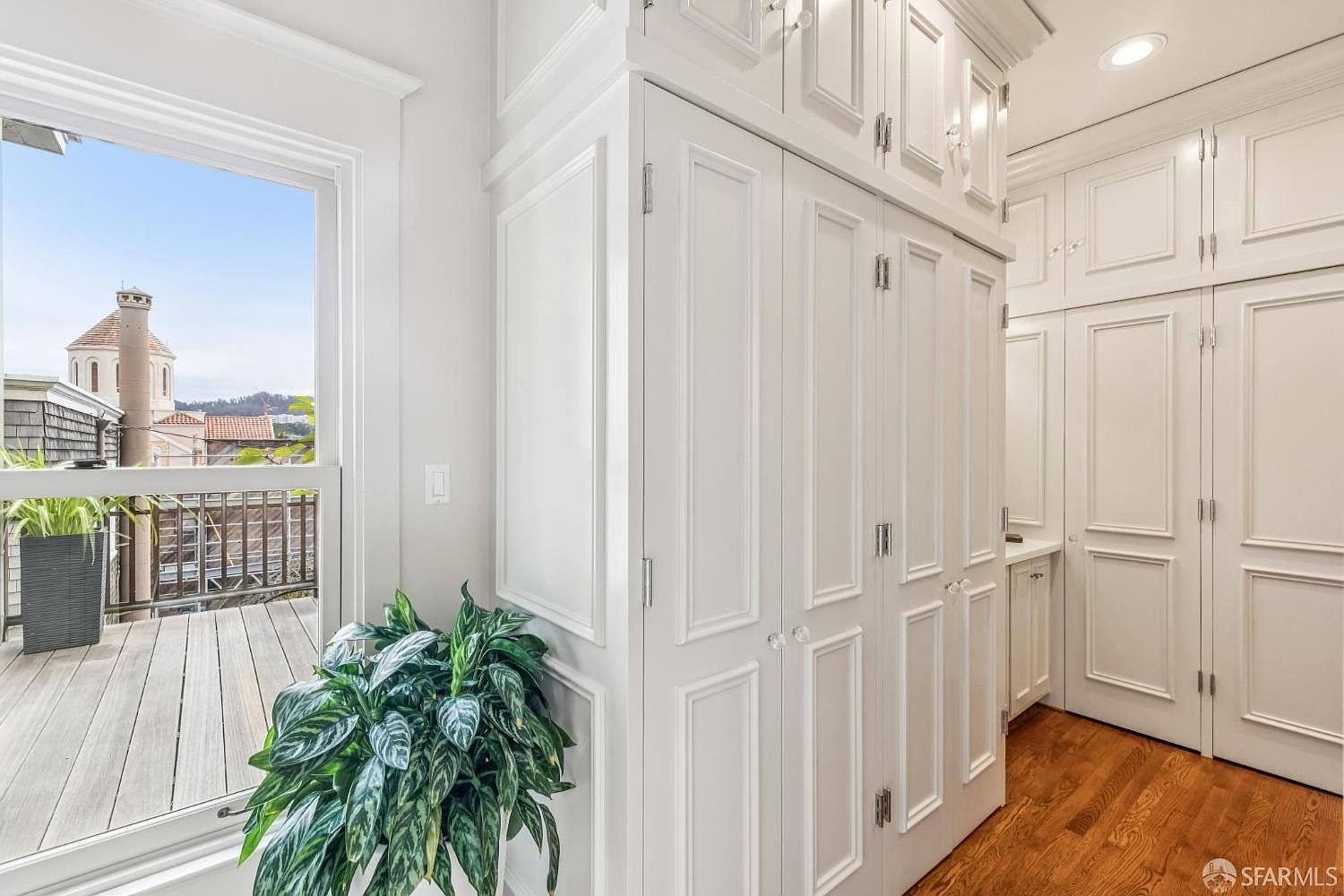 This image showcases a well-organized walk-in closet featuring custom white cabinetry with paneled doors and crystal knobs. The closet is spacious, with ample storage above and below, and the hardwood flooring adds a touch of elegance. A window provides natural light and a view of the outdoors.