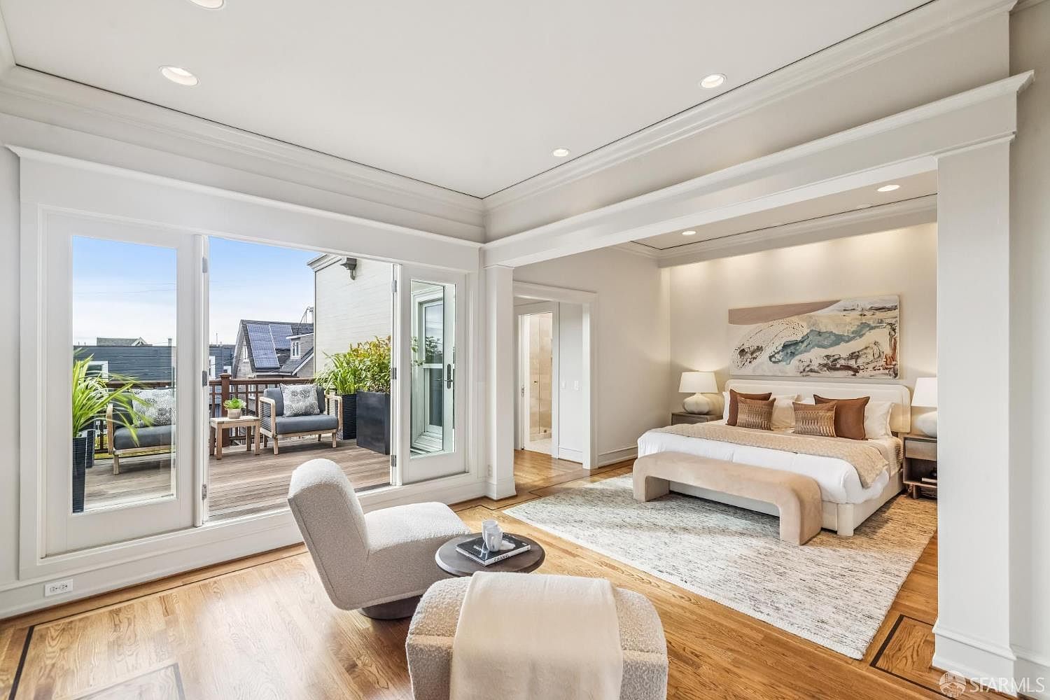 This is a bright and airy primary bedroom featuring a king-size bed with neutral bedding and decorative pillows, complemented by a beige bench. The room opens onto a private balcony with outdoor seating, accessible through large glass doors, and includes a seating area with a modern chair and ottoman. The hardwood floors and neutral color palette create a serene and inviting atmosphere.