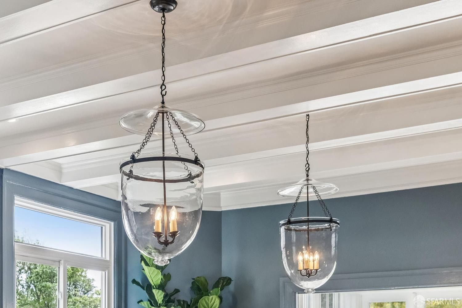The image showcases two elegant glass pendant lights hanging from a white, beamed ceiling. The lights feature a clear glass shade with a black metal frame and chain, creating a sophisticated and modern aesthetic. The room is painted a muted blue, and a window provides natural light, highlighting the fixtures' design.