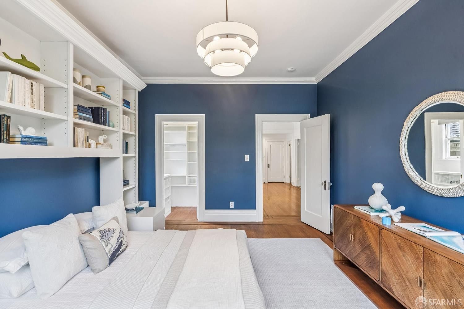 This is a primary bedroom featuring a built-in bookshelf headboard, a modern chandelier, and a blue accent wall. The room has hardwood floors and white trim, with doorways leading to a closet and hallway. A dresser with a round mirror sits against the wall, adding to the room's sophisticated and comfortable ambiance.