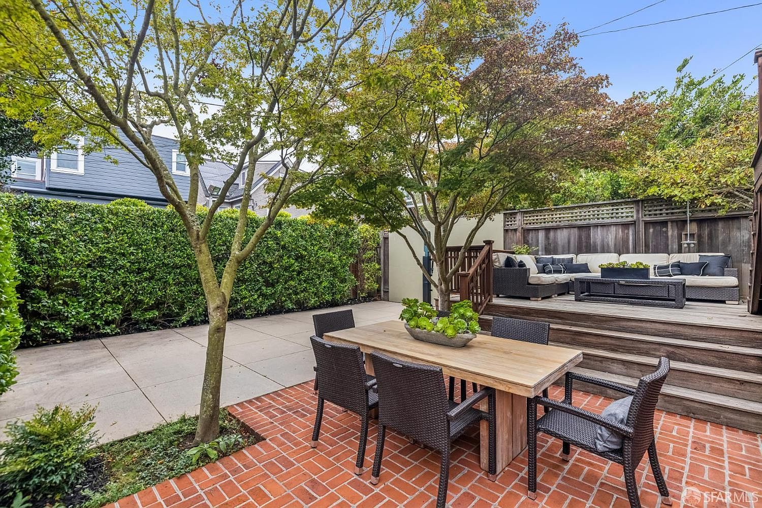 This is a well-maintained backyard featuring a brick patio with a wooden dining table and chairs, a raised wooden deck with outdoor seating, and lush green hedges providing privacy. Mature trees offer shade and add to the serene atmosphere, making it an ideal space for outdoor entertaining and relaxation. The perspective is from ground level, showcasing the depth and layout of the yard.