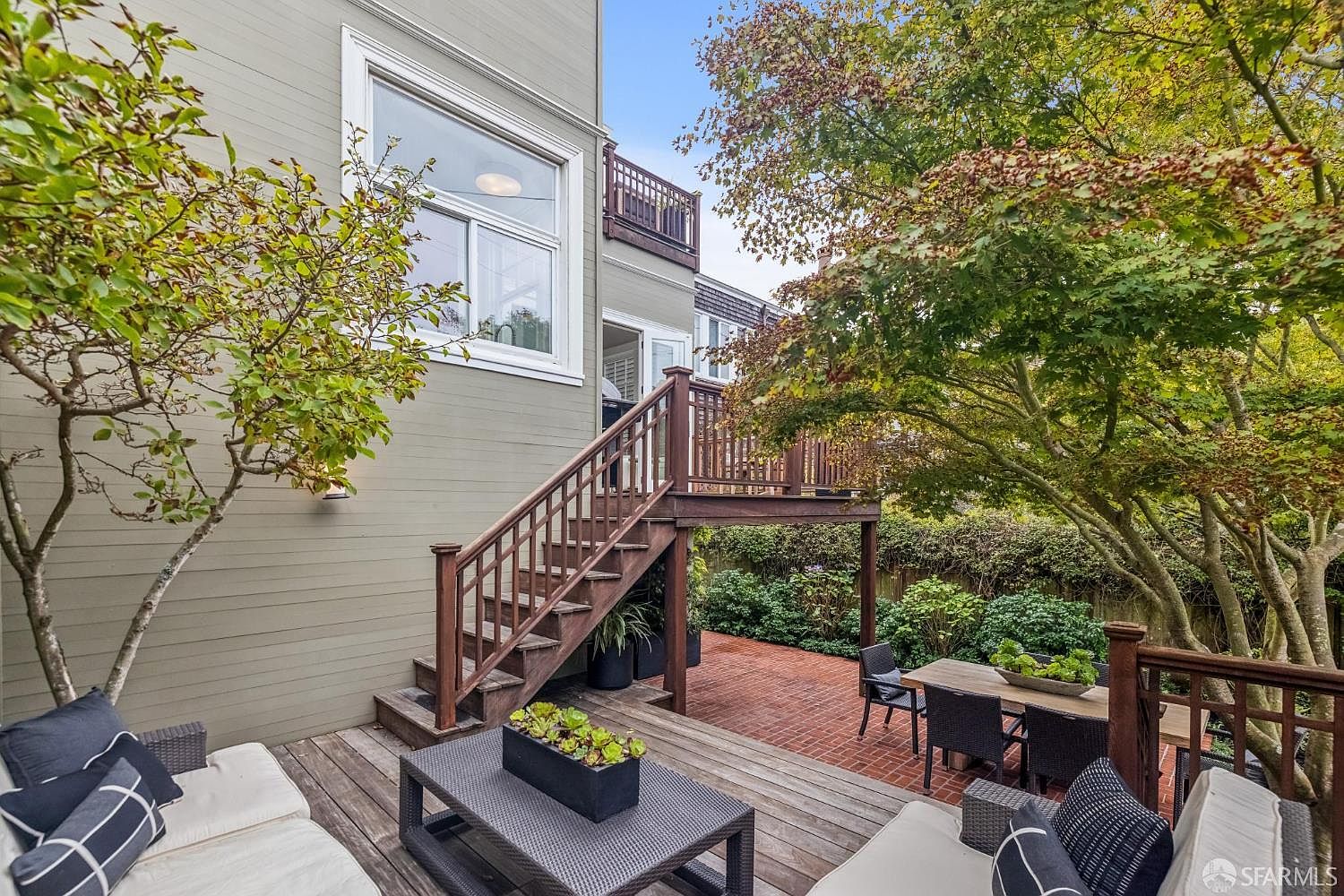 This image showcases a charming outdoor living space with a wooden deck and patio area. A set of stairs leads to an upper deck, while comfortable seating arrangements invite relaxation. The space is surrounded by lush greenery, creating a private and serene atmosphere perfect for entertaining or unwinding.