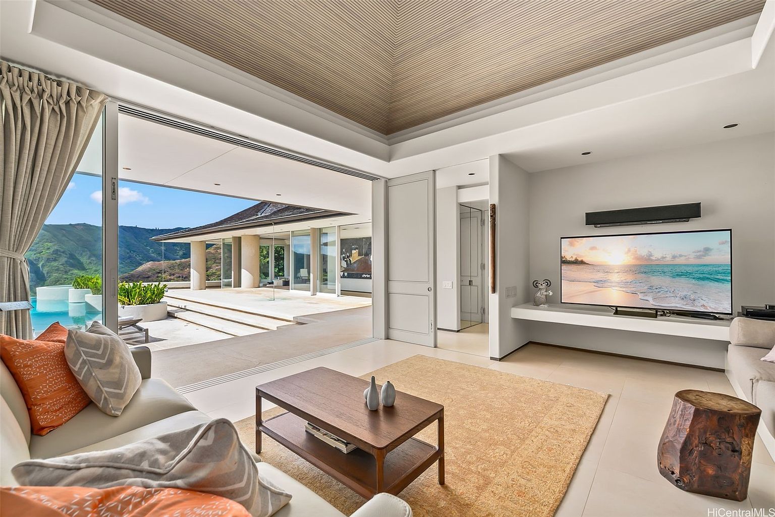 This is a luxurious living room featuring an open floor plan with seamless indoor-outdoor living. The room boasts a modern design with a large sliding glass door leading to a patio and pool area with mountain views. The interior includes a comfortable sofa, a wooden coffee table, a large television, and a unique wooden side table, all contributing to a sophisticated and inviting atmosphere.