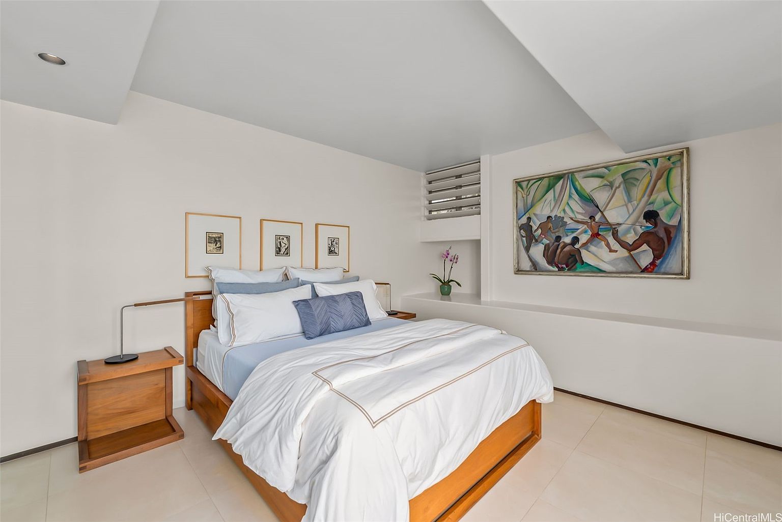 This is a well-lit primary bedroom featuring a wooden bed frame with white linens and blue accent pillows. The room is decorated with framed artwork above the bed and a large painting on the adjacent wall. A wooden nightstand with a lamp sits beside the bed, and the flooring appears to be tiled.