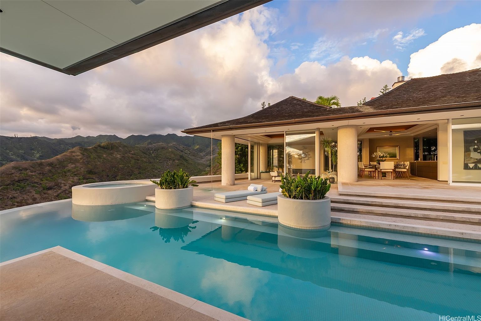 This image showcases a luxurious infinity pool and spa area of a high-end property, with a stunning mountain backdrop. The pool features light blue tiling and is surrounded by a stone patio, complemented by modern lounge chairs and potted plants. The architecture of the house is visible, featuring large glass doors and a covered patio area, creating a seamless indoor-outdoor living experience.