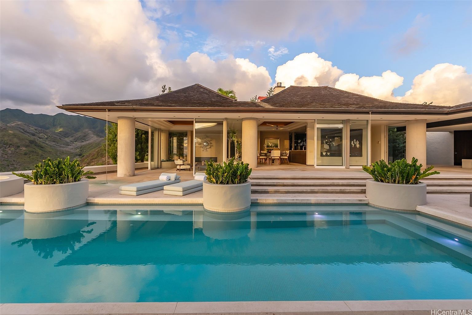 This image showcases a luxurious pool area of a modern home. The pool features clear blue water and is surrounded by a patio with potted plants and lounge chairs. The house has large glass doors that open to the pool area, offering seamless indoor-outdoor living with a mountain view in the background.